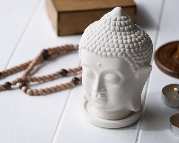 White Buddha head sculpture on a white surface with a wooden box and beads in the background