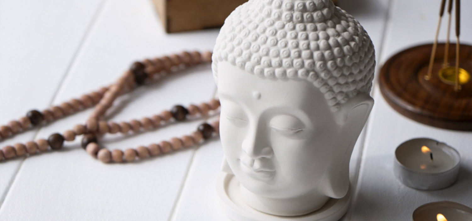 White ceramic Buddha head on a white surface with a candle and beads in the background