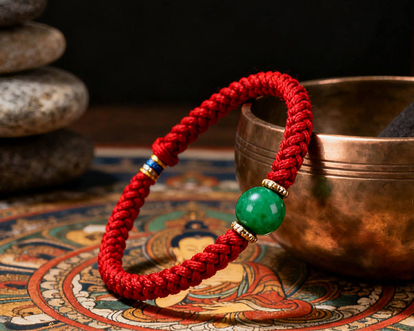 Red braided bracelet with a green bead on a decorative mat with a copper bowl in the background.